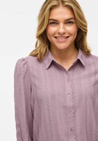 Lys lilla bluse med et teksturert mønster og delikat broderi, knappefront og spiss krage. Lange ermer med rynkede detaljer.