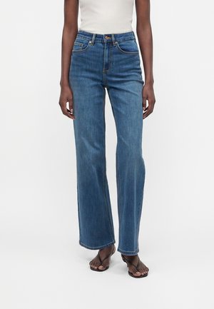 Jean bleu taille haute à jambes droites porté avec des sandales à talons noires à bout ouvert et un haut blanc sans manches.