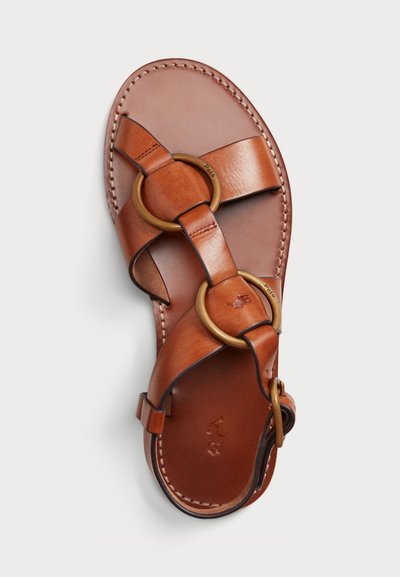 Brūni ādas sandales ar krustveida jostām, kas greznotas ar apaļiem metāla gredzeniem. Tekstūrēta iekšpuse un sānu sprādze, lai pielāgotu izmēru.