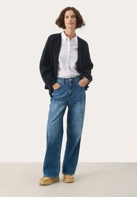 Cardigan lungo nero in maglia sopra una camicia bianca con bottoni, abbinato a jeans blu a vita alta e scarpe slip-on marrone chiaro. Look semplice e informale.