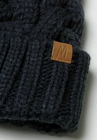 Bonnet tricoté sombre avec une texture côtelée. Une petite étiquette en cuir marron avec un "M" embossé est attachée au revers.
