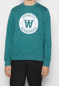 Ciánová svetr s kulatým bílým logem s nápisy "DOUBLE A" a "WOOD WOOD", vyrobený z měkké, texturované látky.