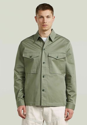 Homme portant une chemise verte claire boutonnée avec deux poches poitrine et un pantalon blanc, debout devant un fond clair.