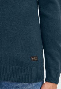 Dunkelblauer Strickpullover mit einem gerippten Saum und Bündchen, versehen mit einem braunen Leder-Label der Marke, das an der unteren Seite angenäht ist. Glatte Textur.