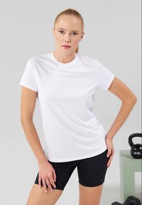 White t-shirt with short sleeves, round neck, and smooth fabric, paired with black shorts. Background features a kettle bell and a green block.