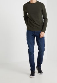 Felpa verde scuro, design aderente, collo rotondo; abbinata a jeans blu. Sneakers nere completano il look casual.