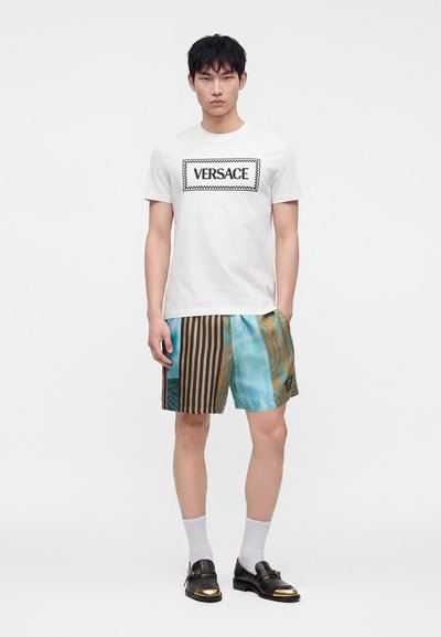 Jeune homme portant un t-shirt blanc Versace, un short rayé bleu et marron à motifs, des chaussettes blanches et des mocassins noirs avec embouts dorés.