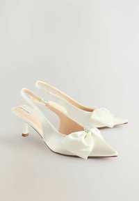 Valkoiset satiiniset slingback-korkokengät, joissa on teräväkärkinen muotoilu ja suurella solki-koristeella edessä, sekä kapea, keskikorkea korko.
