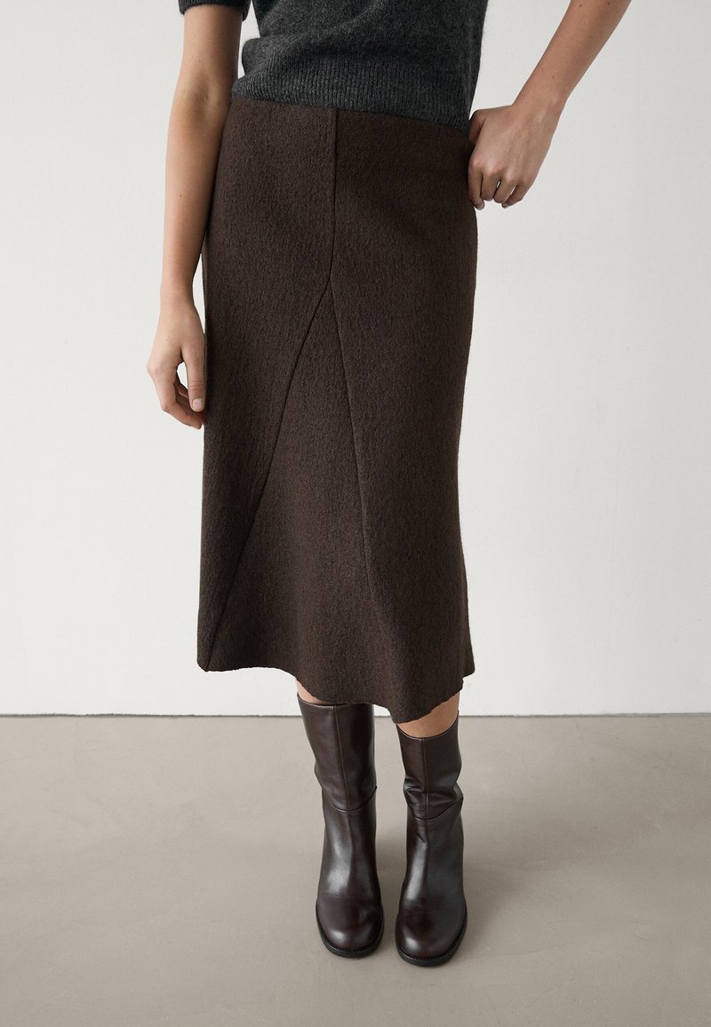 Femme portant une jupe en laine marron foncé longueur genoux, un top à manches courtes gris foncé et des bottes en cuir marron foncé mi-mollet, debout sur un sol uni.