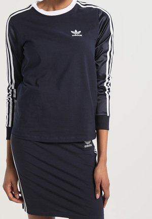 Kobieta ubrana w granatowy, długi top Adidas z długim rękawem oraz pasującą spódnicę z białymi paskami na rękawach i bokach, logo Adidas na piersi i spódnicy.
