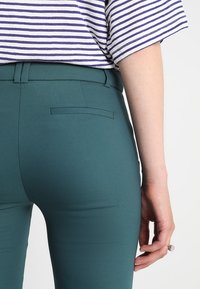 Koppar högmidjade byxor i teal med en slät yta och bakfickor, tillsammans med en vit och lila randig topp.