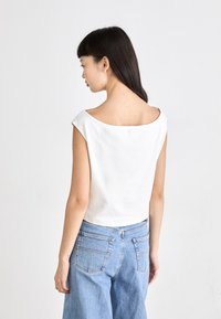 Valge lühike topp, millel on lai, õlgadelt allavõetav kaelus ja lühikesed varrukad. Kombineeritud kõrge vöökohaga helesiniste denim-teksadega. Sile tekstuur.