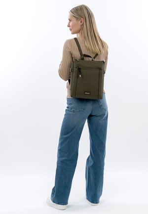 Frau im Profil, die einen beigen Pullover, blaue Jeans, weiße Sneaker trägt und einen olivgrünen rechteckigen Rucksack auf dem Rücken hat.