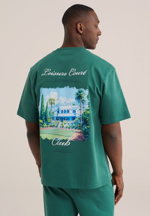 Muškarac u zelenoj majici s šarenom ilustracijom teniskog igrališta, palmi i zgrade s natpisom "Leisure Court Club" na leđima.