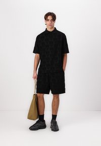Svart kortermet polo skjorte og shorts med et subtilt geometrisk mønster, kombinert med en beige tote-bag og mørke treningssko.