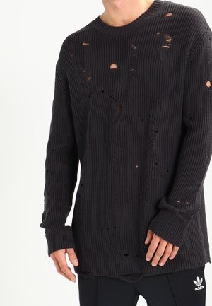 Mand iført en sort oversize striktrøje med flere bevidste huller, kombineret med sorte Adidas-bukser.