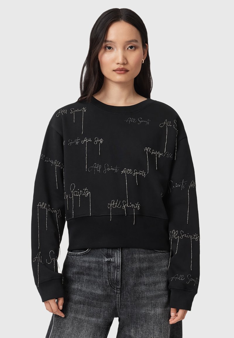 Felpa nera con testo "All Saints" ricamato in argento, colletto a girocollo, vestibilità rilassata e maniche lunghe, abbinata a jeans in denim sbiaditi.