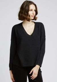 Pull noir en maille avec un col en V, épaules tombantes et poignets côtelés. Tissu doux et texturé, associé à un pantalon noir.