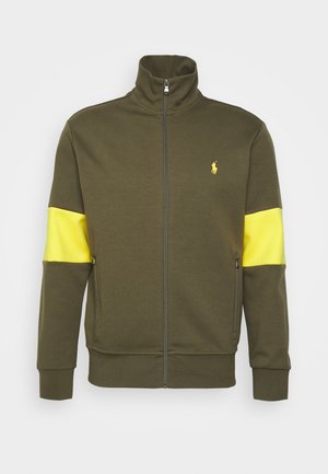 Kurtka na zamek w oliwkowym zielonym kolorze z wysokim kołnierzem, żółtymi paskami na rękawach, kieszeniami na zamek po bokach oraz małym żółtym logo Polo na piersi.