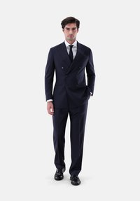 Mann steht, trägt einen marineblauen zweireihigen Anzug mit weißem Hemd, schwarzer Krawatte und schwarzen Anzugschuhen, vor einem schlichten weißen Hintergrund.