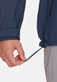 Giacca blu navy con finitura strutturata; mano che regola il cordino in vita; pantaloni grigi chiari visibili nella parte inferiore dell'immagine.