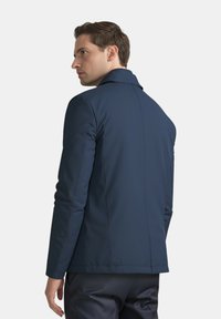 Uomo che indossa un blazer blu navy aderente, visto di spalle e leggermente girato di lato su uno sfondo chiaro e semplice.