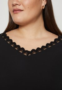 Crna majica s detaljem od čipke na izrezu. Glatka tkanina s V-izrezom. Približan pogled na izrez i gornji dio prsnog koša.