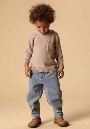 Malé dítě s kudrnatými vlasy, oblečené v béžovém svetru, světle modrých džínách a hnědých botách, stojící před jednoduchým béžovým pozadím.