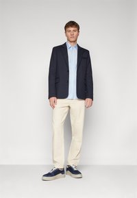 Navy blazer over a light blue striped shirt, cream trousers, and navy sneakers with a textured sole. Model standing against a plain background.