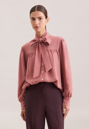 Lichtroze blouse met een grote strik kraag, knopen aan de voorkant en gather-details bij de schouders. Soepele stof met luchtige mouwen.