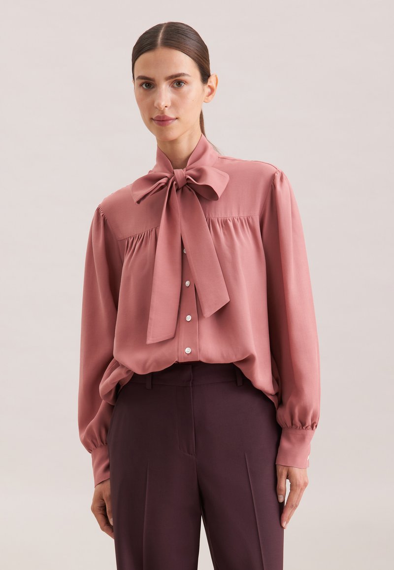Blusa rosa chiaro con un ampio colletto a fiocco, chiusura frontale a bottoni e dettagli arricciati sulle spalle. Tessuto morbido con maniche svasate.