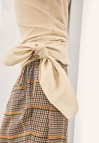 Haut beige léger noué à la taille, associé à un pantalon à carreaux avec des lignes marron, vertes et orange, mettant en valeur les rayures verticales.