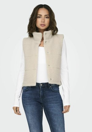 Beige polár mellény magas nyakkal, kapcsoló gombokkal és doboz alakú dizájnnal. Fehér hosszú ujjú inggel és sötét farmernadrággal párosítva.