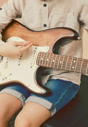 Enfant portant une chemise beige et un short en jean, jouant d'une guitare électrique marron et blanche en étant assis.