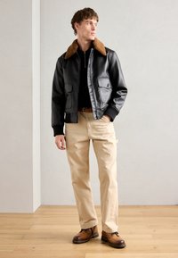 Black leather bomber jacket with brown fur collar, paired with beige chinos and brown lace-up boots. Simple, structured design, and fitted look.