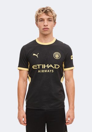 Jeune homme debout, faisant face, portant un maillot de football noir de Manchester City avec des bordures jaunes et des logos de sponsors.