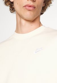 Světlé krémové bavlněné tričko s kulatým výstřihem a krátkými rukávy. Na hrudi je malé vyšívané bílé logo Nike.