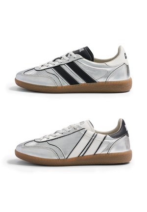 Deux paires de baskets en cuir argenté avec semelles en gomme, l'une avec des rayures noires et une languette noire, l'autre avec des rayures blanches et une languette blanche, vues de côté.