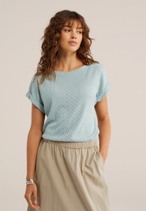 Frau mit welligem braunem Haar, die ein hellblaues, strukturiertes kurzärmeliges Oberteil trägt, das in einen beigen Rock mit elastischem Bund gesteckt ist, und mit einer Hand in der Tasche steht.