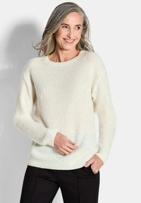 Strickpullover in Creme mit Rautenmuster, Rundhalsausschnitt, gerippten Bündchen und Saum. Getragen mit schwarzen Hosen, die eine strukturierte Oberfläche aufweisen.