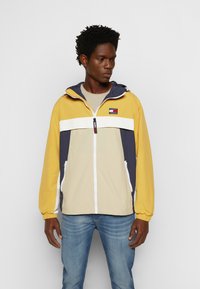Giacca a vento con corpo giallo, dettagli blu navy e striscia orizzontale bianca. Presenta un cappuccio, una cerniera frontale e due tasche laterali.