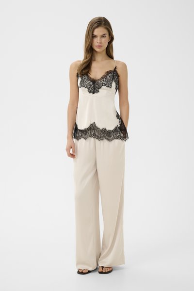 Femme portant une camisole en satin crème avec une bordure en dentelle noire et un pantalon large assorti en satin, debout devant un fond blanc uni.