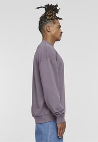Felpa a girocollo viola con maniche lunghe, realizzata in morbido tessuto. Polsini e orlo a coste, abbinata a jeans di denim azzurro chiaro.