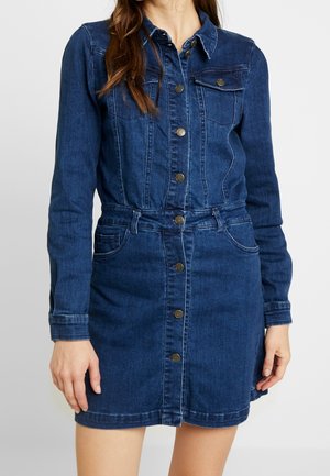 Kobieta ubrana w ciemnoniebieską sukienkę z denimu z długimi rękawami i kieszeniami z przodu, stojąca na tle jednolitego jasnego tła.