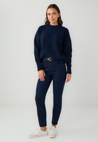 Donna in piedi con un maglione blu navy e pantaloni, indossando sneakers bianche, su uno sfondo chiaro e semplice.