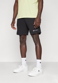 Black cotton shorts with a relaxed fit, featuring a small white and red Champion logo. Worn with white sneakers and socks.