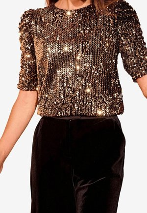 Femme portant un haut bronze à paillettes à manches courtes associé à un pantalon en velours noir taille haute, torse visible avec les bras étendus.