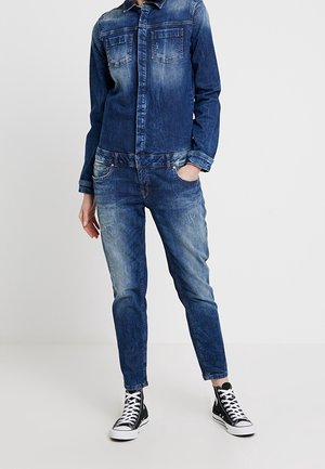 Personne portant une chemise en denim bleu délavé et un jean, avec des baskets hautes noires et blanches, debout avec une main dans la poche.