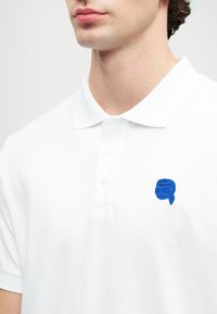 Muž v bielom polo tričku s malým modrým vyšívaným logom tváre s okuliarmi a prilbou na ľavej strane hrude.