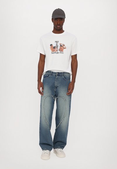 Jeune homme portant un t-shirt blanc à motif graphique, un jean bleu avec des déchirures en forme de cœur, des baskets blanches et une casquette de baseball grise, debout sur un fond blanc.